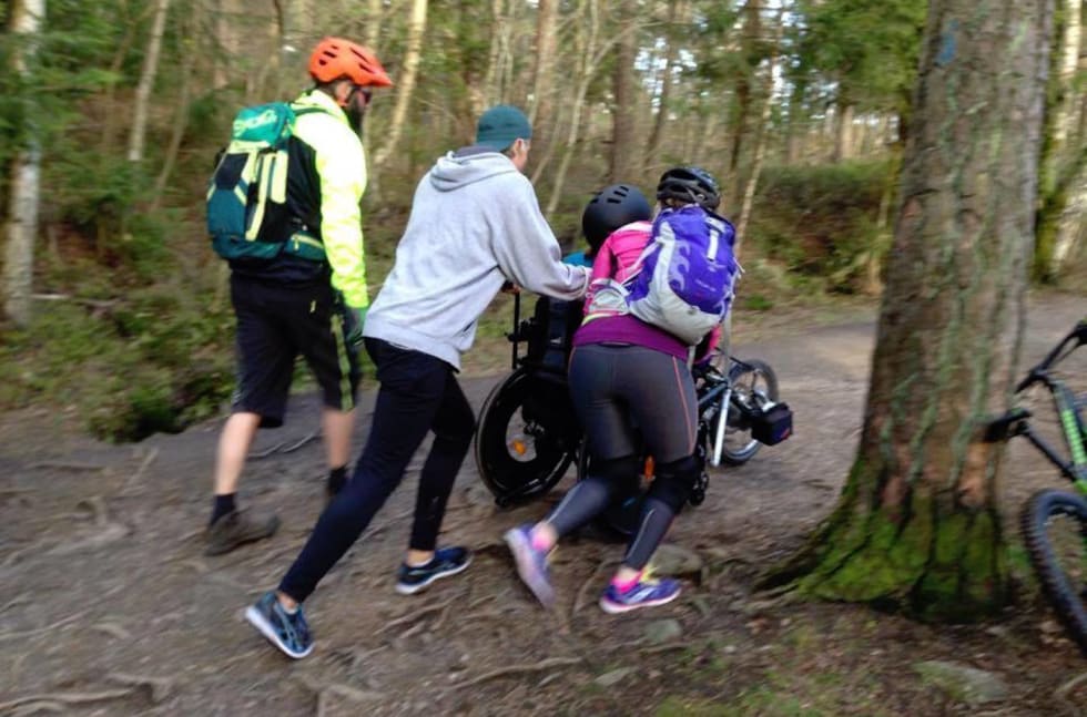En strategisk hjelpende hånd eller fem kommer godt med når en skal over røtter og crux. Foto: Gresvik IF Stisyklister GIF Handicapsykling strategisk hjelp- Foto GIF stisyklister 1400x924